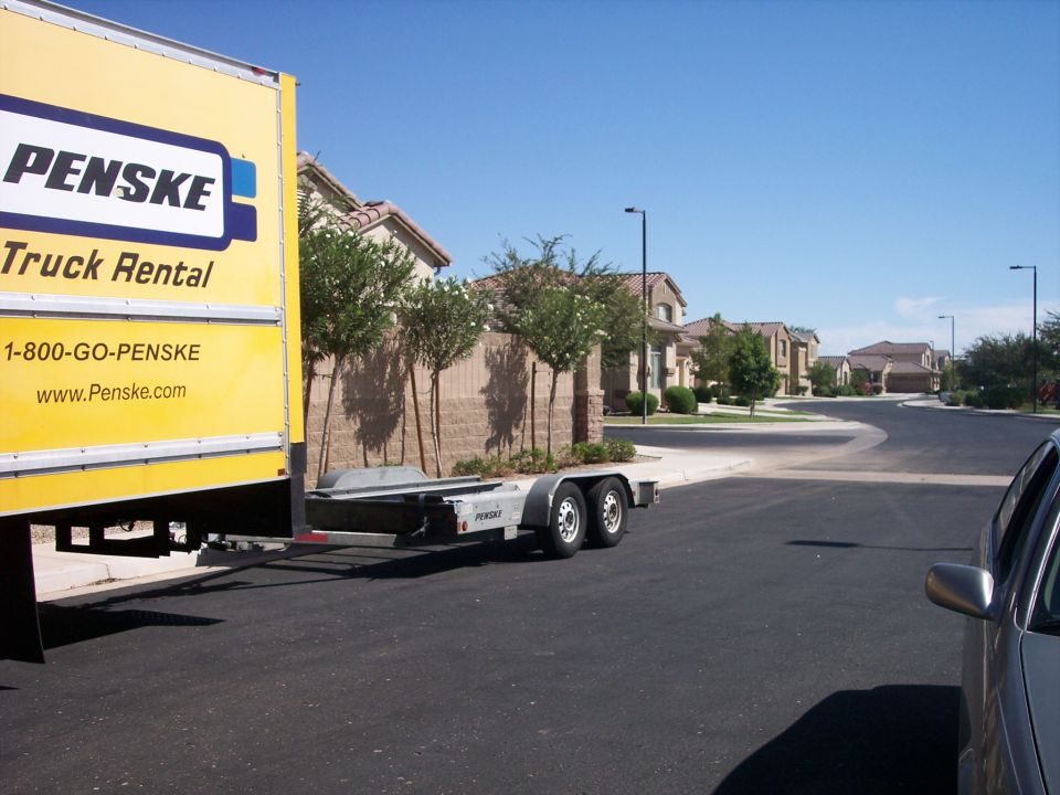Yellow Penske box truck with empty tandem-axle car trailer on a suburban street