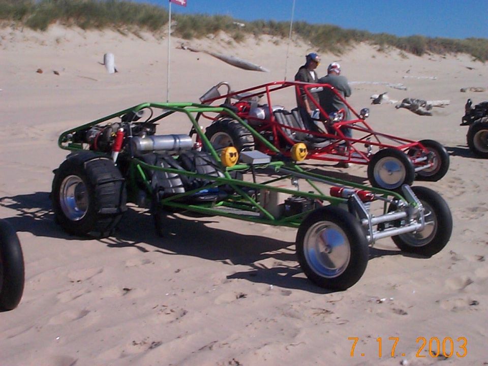 Green and red tube-frame dune buggies parked on sand with riders and ATV nearby