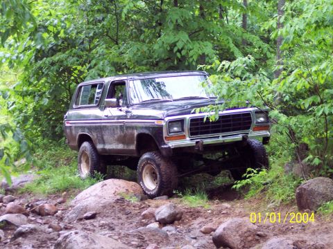 1979 Ford Bronco - Black Ford Bronco driving off-road through a wooded, rocky area.