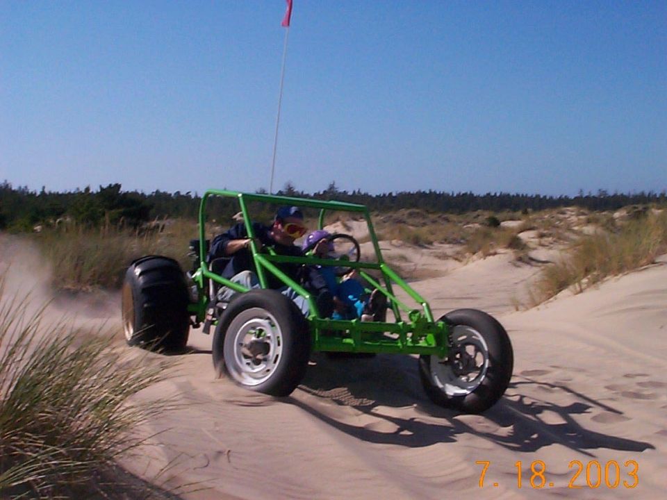 Green tube-frame dune buggy with riders driving across sand dunes