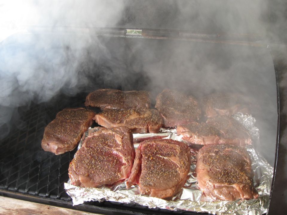 Several raw steaks seasoned and cooking on a grill with smoke rising