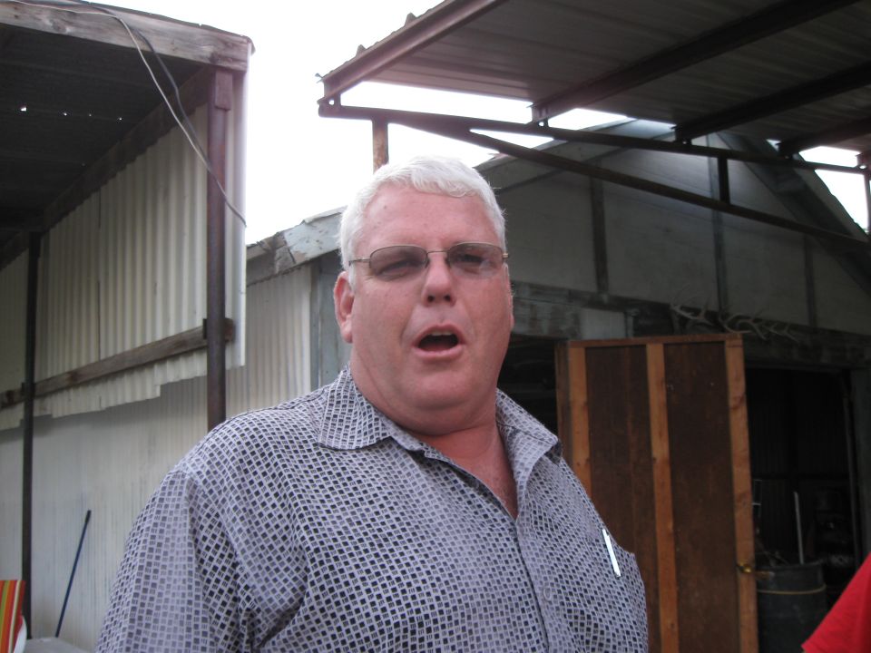 Man with white hair and glasses, mouth open, wearing a patterned shirt. Behind him are corrugated metal buildings