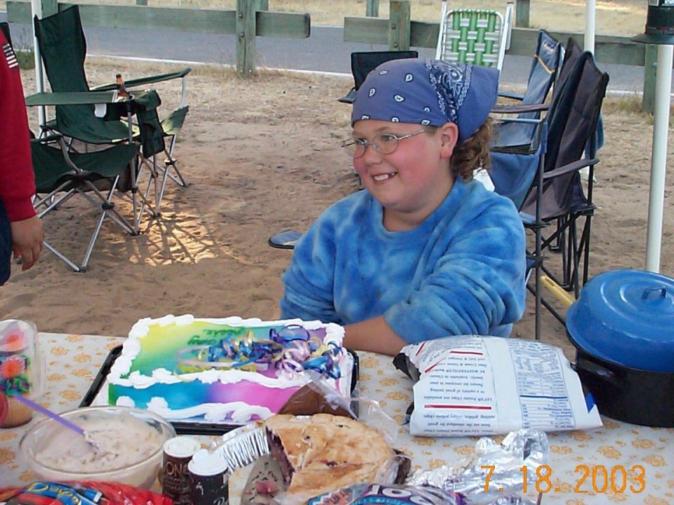 Person in a blue bandana at a campsite picnic table with a rainbow birthday cake