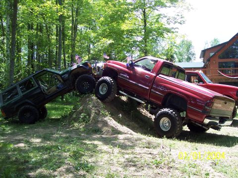 1979 Ford Bronco - Two modified trucks tackle an off-road course with a house in the background.