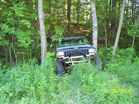 1979 Ford Bronco - A Jeep Cherokee XJ navigates a wooded trail, its front end visible through tall grass and trees.