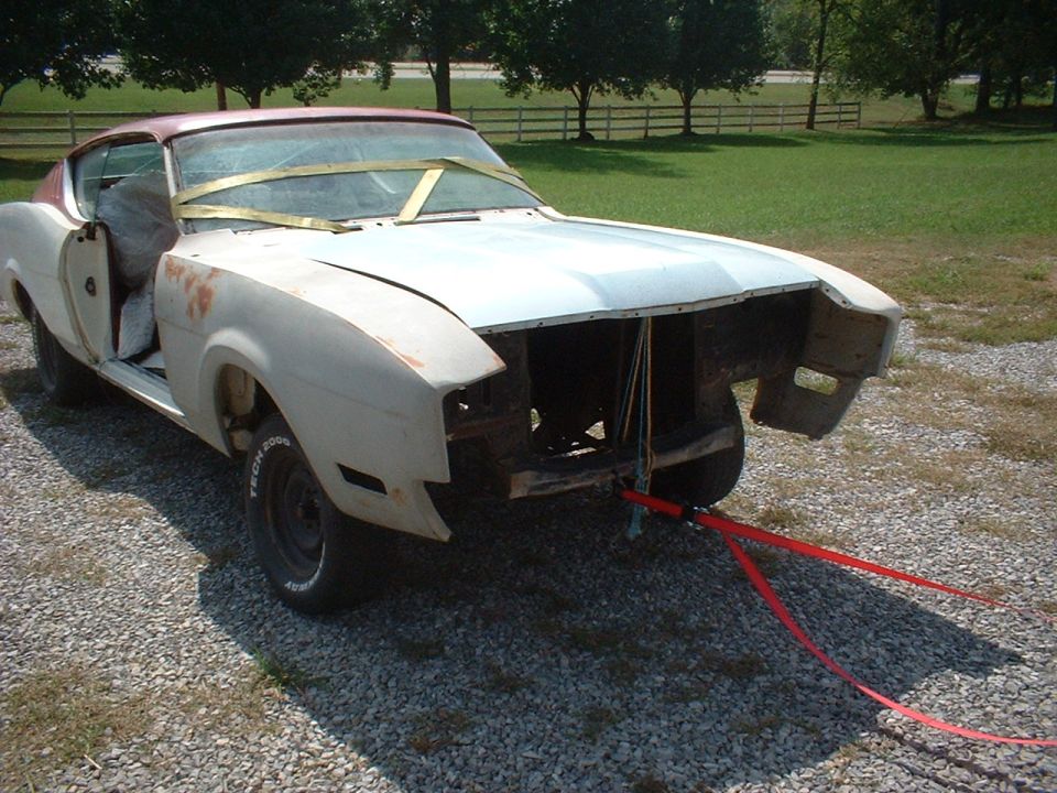 Stripped primer-white fastback muscle car project on gravel with tow strap and no front end