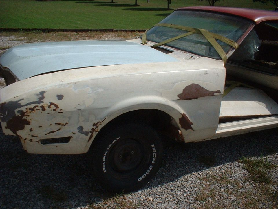 Classic coupe in primer with rust patches mid-restoration, front three-quarter view on gravel