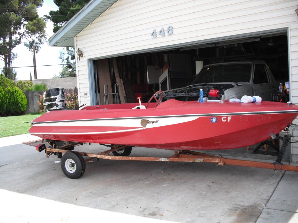Red boat with a white stripe on a rusty trailer, parked in front of a garage with a truck inside