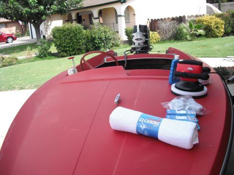 1991 Ford Bronco - A red boat is prepared for cleaning with a polisher and supplies laid out on its deck, while a red car is parked in the driveway behind it.