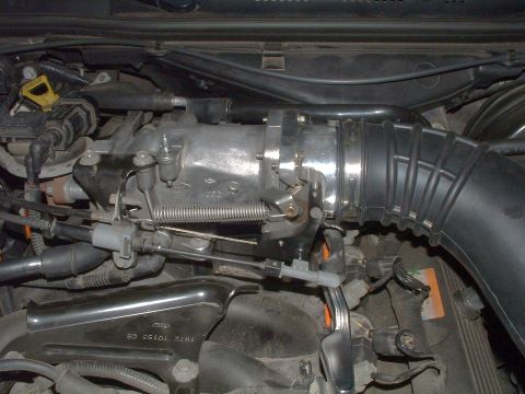 2004 Ford Crown Victoria - Under-hood view of a V8 intake area showing the cast-aluminum throttle body and elbow with Ford oval casting mark, a coiled throttle return…