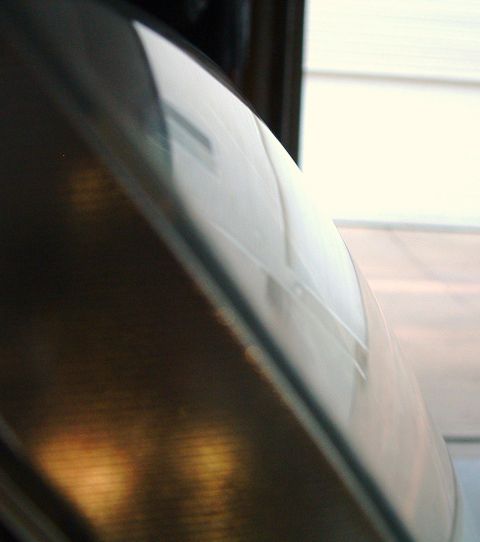 2004 Ford Crown Victoria - Tight edge-on close-up of a headlight lens surface catching daylight, with warm amber reflections along the lower reflector and a white hou…