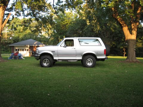 1994 Ford Bronco - A silver Ford Bronco is parked on a grassy lawn in front of a house and trees.