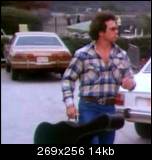 1978 Ford Mustang II - A man carrying a guitar case walks past a brown sedan in a parking lot.