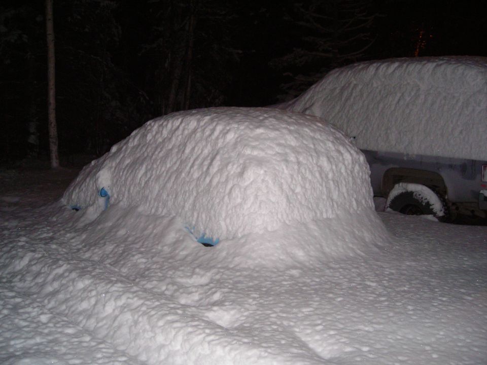 Two vehicles buried under thick snowdrifts, with blue plastic visible through openings in the snow on the closest vehicle