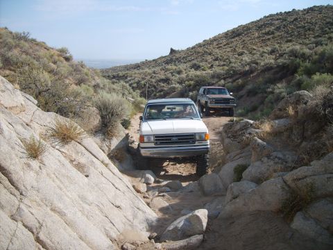 1989 Ford Bronco - A white 1987 Ford Bronco navigates a challenging rocky off-road path, with a second SUV following closely behind.