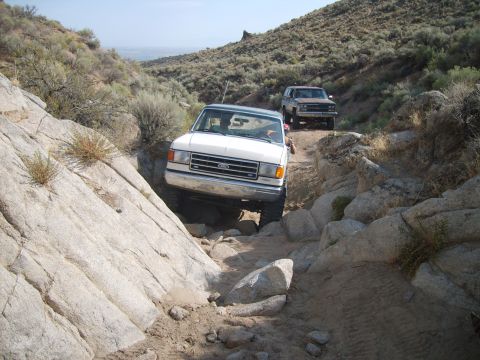 1989 Ford Bronco - Two trucks tackle a challenging off-road path in a desert landscape.