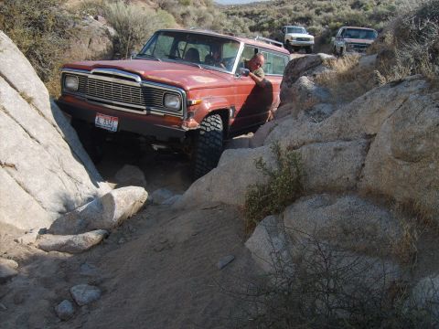 1989 Ford Bronco - A red Jeep Wagoneer is shown off-roading on a rocky trail, with two other vehicles following.