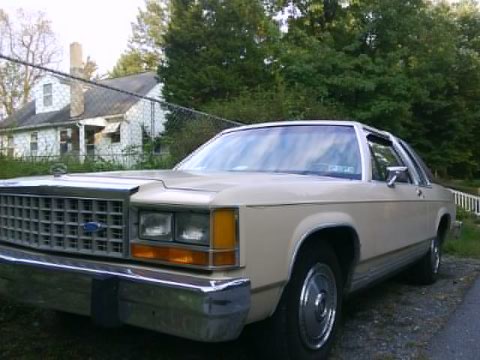 1986 Ford Crown Victoria LTD - A tan 1986 Ford Crown Victoria LTD sedan is parked in front of a house with trees in the background.