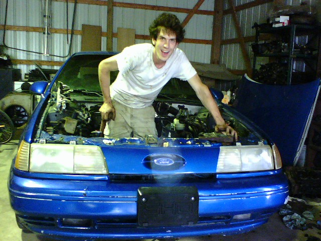 Young man leaning over open engine bay of blue Ford Tempo or Topaz in shop