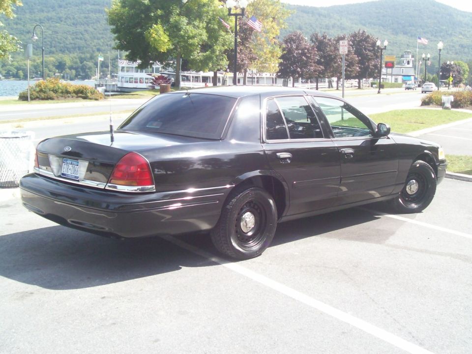 Black Ford Crown Victoria sedan with black steel wheels, rear three-quarter view, parked near a lake