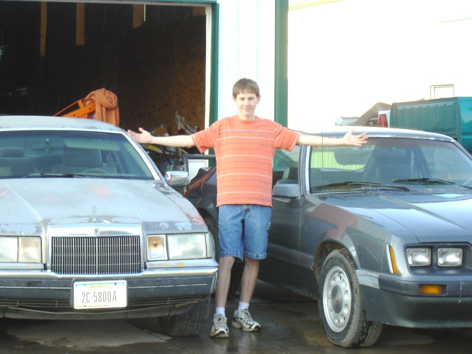 Boy with arms spread between a silver sedan and a gray Fox-body Mustang outside a garage