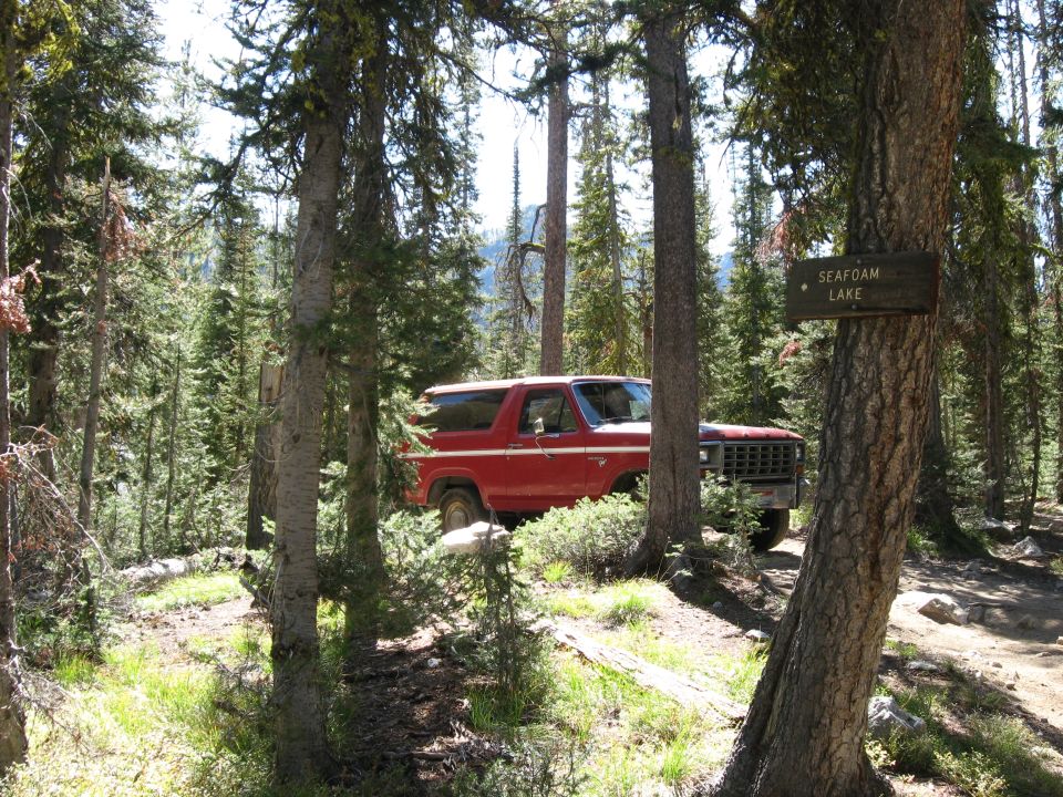 Red Ford Bronco parked on a dirt path in a forest, with a sign pointing to Seafoam Lake