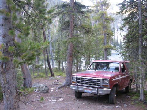 1981 Ford Bronco - A red SUV is parked in a wooded area, with a lake visible through the trees.