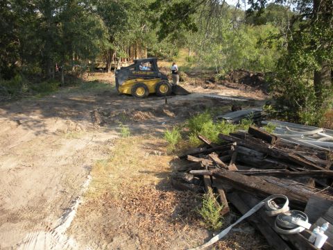 1996 Ford Bronco - A yellow skid steer loader is parked in a clearing surrounded by trees, with a pile of lumber in the foreground.