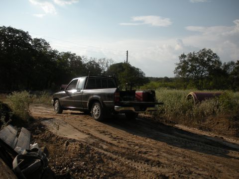 1996 Ford Bronco - A dark pickup truck navigates a muddy dirt road through a rural landscape under a partly cloudy sky.