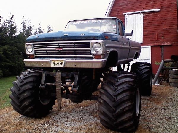 Lifted blue Ford monster truck with huge tires parked by a red barn