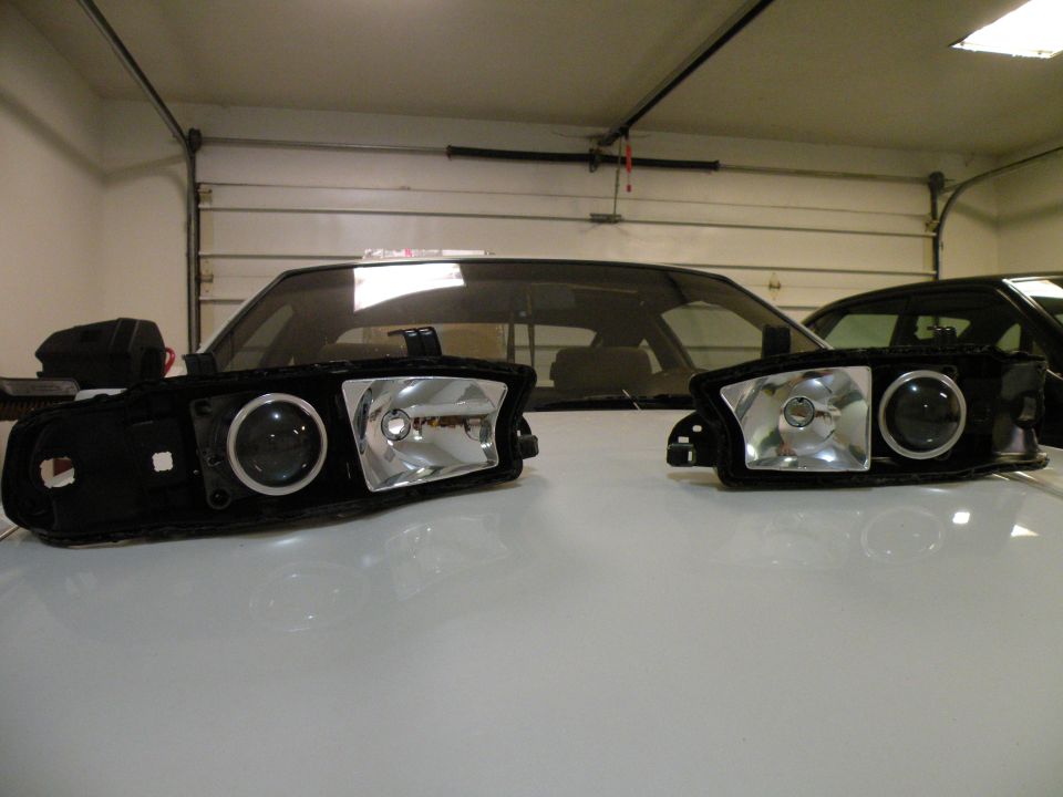 Two aftermarket projector headlights sit on a white surface in front of a car's windshield and a garage door