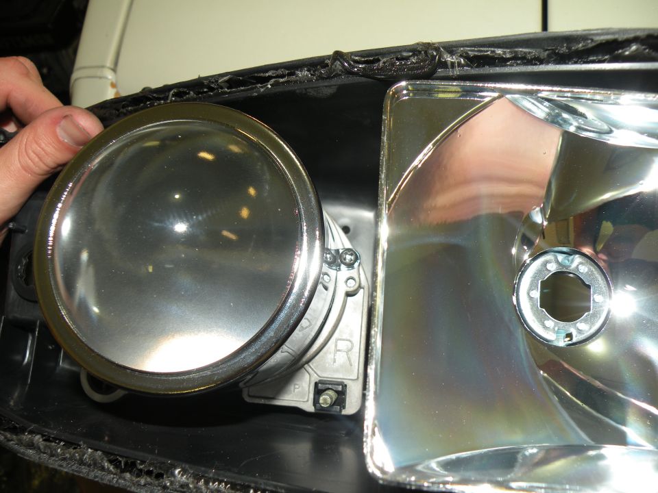 Close-up of a car headlight assembly with a hand holding a projector lens. The housing shows a reflective surface and a part labeled 'R'
