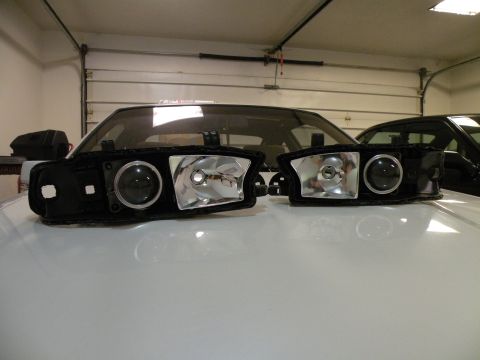 1998 Subaru Legacy - A pair of aftermarket projector headlights are displayed in a garage, with the hoods of two vehicles visible in the background.