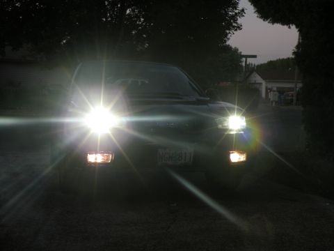 1998 Subaru Legacy - The front of a dark-colored vehicle is illuminated by its bright headlights at dusk.