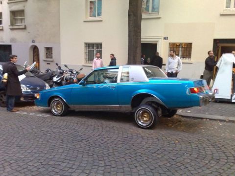 1991 Ford Crown Victoria - A blue lowrider with gold rims is parked on a cobblestone street, with people and motorcycles nearby.