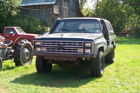 1988 Chevrolet Blazer - A black Chevrolet K5 Blazer with off-road modifications sits in a grassy yard next to a red pickup truck and a tractor.