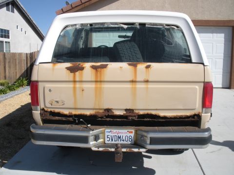 1990 Ford Bronco - Rear view of a 1990 Ford Bronco with extensive rust damage on the lower body and tailgate.