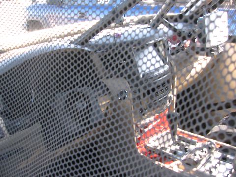 1990 Ford Bronco - close-up shot through a perforated metal screen revealing chrome mechanical components behind it.
