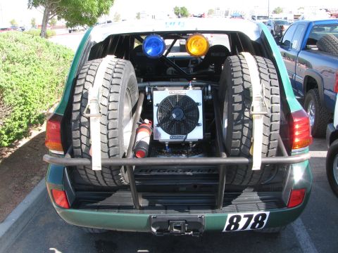 1990 Ford Bronco - open rear of a green off-road race SUV shows twin spare tires, a mounted radiator fan and race number 878.