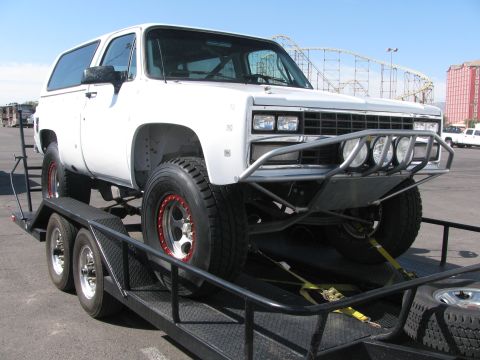 1990 Ford Bronco - white lifted Chevrolet Blazer fitted with an off-road brush guard sits strapped on a trailer in a paved lot.