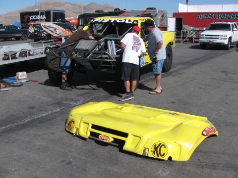 1990 Ford Bronco - Mechanics work on a yellow off-road race truck in a pit area, its yellow KC-stickered body panel lying on the pavement.
