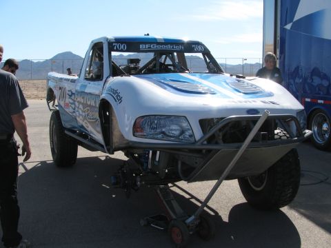 1990 Ford Bronco - white-and-blue Ford off-road race truck numbered 700 sits raised on a transmission jack at a desert event.