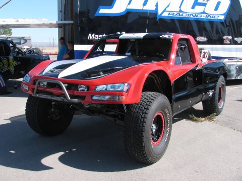 1990 Ford Bronco - red-and-black off-road race truck numbered 30 with long-travel front suspension parked before a Jimco Racing trailer.