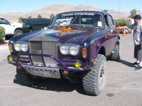 1990 Ford Bronco - purple Rolls-Royce-bodied car mounted on a lifted off-road chassis with big tires and Think Strange windshield banner.