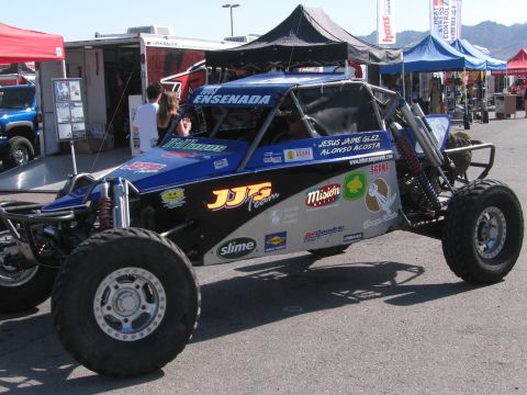1990 Ford Bronco - blue open-cockpit tube-frame off-road race buggy with sponsor decals and big knobby tires parked among canopy tents.