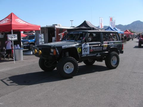 1990 Ford Bronco - black lifted Jeep Cherokee race vehicle numbered 1701 with a front brush guard and yellow auxiliary lights in a desert pit.