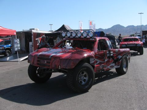 1990 Ford Bronco - red and silver graphically wrapped off-road race truck with a roof-mounted light bar parked in a paved desert pit area.