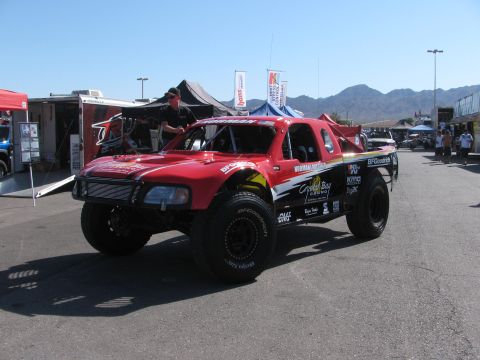 1990 Ford Bronco - red off-road race truck with black wheels and multiple sponsor decals being prepped in a sunny pit area.