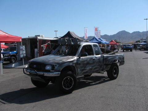 1990 Ford Bronco - gray-camo Toyota Tacoma prerunner race truck numbered 741 with overhead lights and a long-travel suspension in a desert pit.