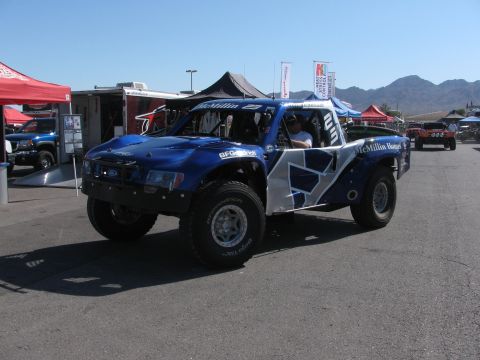 1990 Ford Bronco - blue and white Ford off-road trophy truck with McMillin Racing graphics and BFGoodrich tires in a sunny pit area.